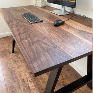 Puede incluir: Un escritorio rectangular con una superficie de madera de color marr&oacute;n oscuro con patrones naturales visibles. El escritorio est&aacute; sostenido por patas de metal negro dispuestas en un marco inclinado. Un teclado y un rat&oacute;n negros est&aacute;n sobre &eacute;l.