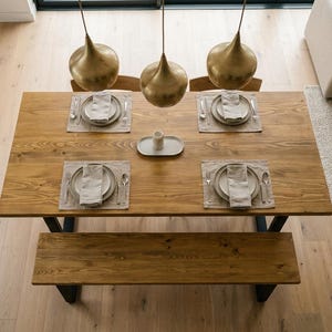 May include: An overhead shot of a wooden dining table set for four. Each place setting has a plate, napkin, and cutlery on a neutral placemat. A bench is below the table. Three gold pendant lights hang above.