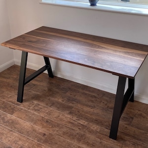 Puede incluir: Un escritorio rectangular con una superficie de madera de color marr&oacute;n oscuro con patrones naturales visibles y un acabado liso. El escritorio se apoya en dos patas de metal negro en forma de A inclinadas hacia afuera para mayor estabilidad.