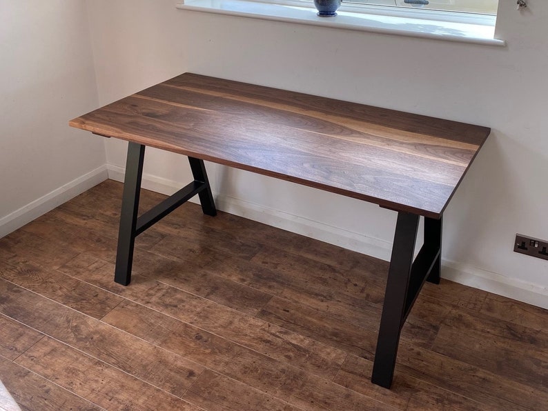 Puede incluir: Un escritorio rectangular con una superficie de madera de color marr&oacute;n oscuro con patrones naturales visibles y un acabado liso. El escritorio se apoya en dos patas de metal negro en forma de A inclinadas hacia afuera para mayor estabilidad.