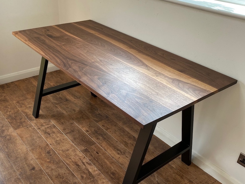 Puede incluir: Un escritorio rectangular con una superficie de madera de nogal marr&oacute;n oscuro con vetas naturales visibles y un acabado liso. El escritorio se apoya sobre dos patas de metal negro en forma de A conectadas por una barra de soporte horizontal.