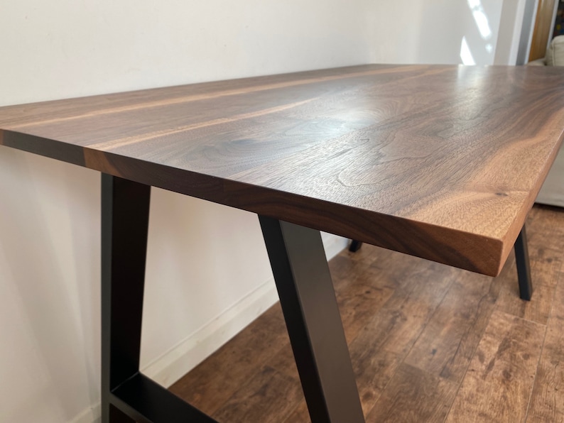 Puede incluir: Una mesa rectangular con una superficie de madera natural de color marr&oacute;n oscuro y veteado visible. La mesa se apoya sobre patas de metal negro mate, robustas y anguladas hacia afuera, que proporcionan una base estable y firme.