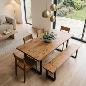 May include: A wooden dining table with a matching bench and four chairs. The table and bench have black metal legs. A vase of greenery sits on the table. The room has a neutral colour palette with a view of the outdoors.