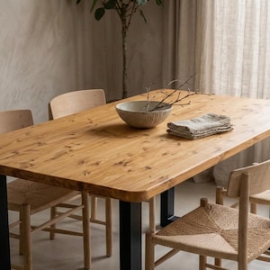 May include: A rectangular dining table with a natural light wood grain top and thick black metal legs, accompanied by four wooden chairs featuring woven seats. A ceramic bowl with twigs and a folded linen napkin sit on the table surface.