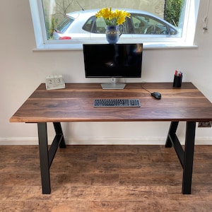 Puede incluir: Un escritorio rectangular con una superficie de madera de color marr&oacute;n oscuro con vetas visibles y patas de metal negro. El escritorio sostiene un monitor de computadora, un teclado, un rat&oacute;n, un calendario de bloques de madera con el d&iacute;a 23 y un portal&aacute;pices negro.