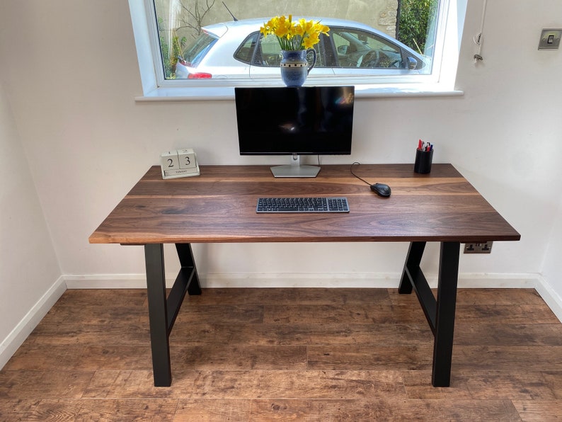 Puede incluir: Un escritorio rectangular con una superficie de madera de color marr&oacute;n oscuro con vetas visibles y patas de metal negro. El escritorio sostiene un monitor de computadora, un teclado, un rat&oacute;n, un calendario de bloques de madera con el d&iacute;a 23 y un portal&aacute;pices negro.