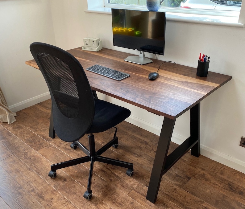 Puede incluir: Un escritorio rectangular de madera oscura con veta natural y patas de metal negro en forma de A. El escritorio se acompa&ntilde;a de una silla de oficina negra de malla con ruedas. Sobre la superficie hay un monitor, un teclado, un rat&oacute;n y un portal&aacute;pices.
