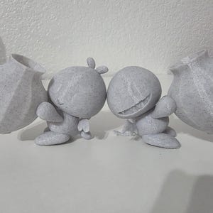 Puede incluir: Cuatro figuras grises, cada una sosteniendo un jarrón texturizado. Las figuras tienen cuerpos redondeados, brazos y piernas pequeños, y expresiones sonrientes o neutras. Los jarrones tienen forma bulbosa con superficie texturizada.