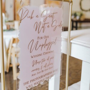 May include: A clear acrylic wedding sign with gold lettering. The sign reads "Pick a Seat Not a Side For Our Unplugged Wedding Ceremony. Please turn off your cameras & cell phones AND JUST BE OUR GUEST. THE PHOTOGRAPHERS HERE WILL HANDLE THE REST."