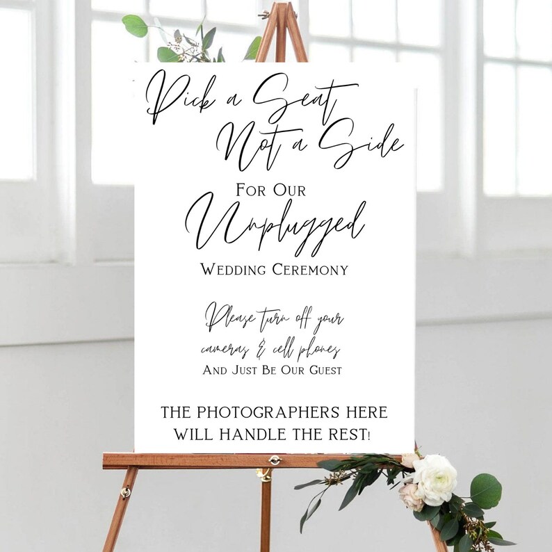 May include: A white sign on a wooden easel with black script. The sign reads "Pick a Seat Not a Side For Our Unplugged Wedding Ceremony." Additional text requests guests turn off cameras and cell phones. Floral accents are visible.