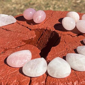 May include: Assortment of polished rose quartz and clear quartz stones in various shapes, including rounded and oval forms. The stones are displayed on a weathered, red-painted wooden surface, with natural sunlight illuminating the crystals.