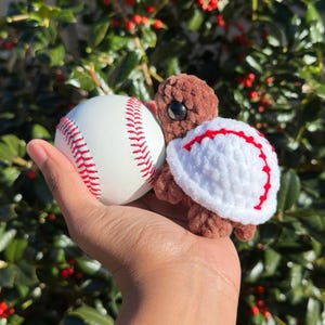 May include: A handmade crochet turtle with a white shell featuring red stitching, next to a white baseball with red stitching. The turtle is brown with large black eyes. The baseball is held in a hand.