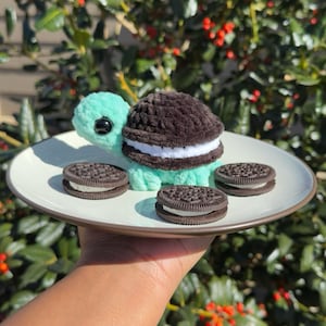 May include: A crocheted turtle plush with a mint green head and limbs, featuring a dark brown and white sandwich cookie-shaped shell. The plush sits on a white plate alongside three round, dark chocolate sandwich cookies with white cream filling.