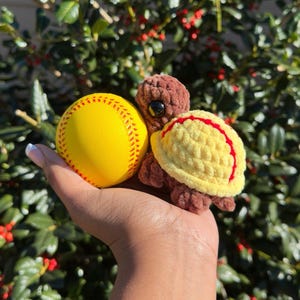 May include: A yellow softball with red stitching next to a brown and yellow crocheted turtle. The turtle has a yellow shell with red trim and large black eyes. The items are held in a hand.
