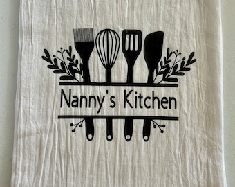 May include: White kitchen towel with black text that reads "Nanny's Kitchen". The text is surrounded by a black silhouette of kitchen utensils and leafy sprigs.