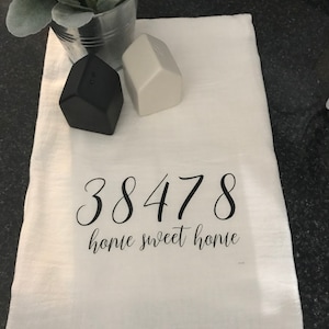 May include: White kitchen towel with black text that reads "38478 home sweet home". Two small house-shaped salt and pepper shakers are on the towel, one black and one white.