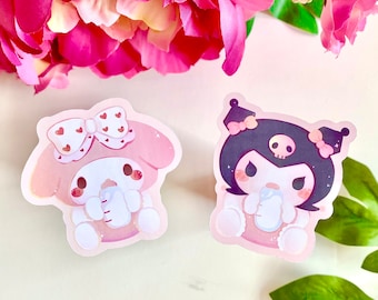 May include: Two cartoon character stickers, one pink and one black, both holding a bottle. The pink character has a pink bow with white hearts and the black character has a black bow with a white skull. Both characters have big eyes and are smiling.