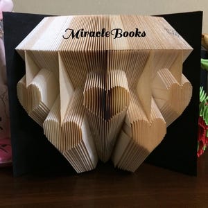 Może przedstawiać: Rzeźba z książki z wieloma kształtami serc utworzonymi ze stron, z napisem Miracle Books wydrukowanym na czarno na górnej krawędzi. Rzeźba znajduje się na jednolitym czarnym tle, prezentując skomplikowane zagięcia papieru.