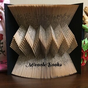 May include: A folded book art sculpture featuring pages intricately creased into a geometric pattern with a flared top and bottom. The centre of the sculpture displays the black text Miracle Books printed directly onto the paper pages.