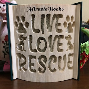 Puede incluir: Un libro tallado con las palabras "LIVE LOVE RESCUE" cortadas en las páginas. El diseño incluye huellas de patas y siluetas de perros. El libro tiene una columna vertebral verde y el texto "Miracle Books" está escrito en la parte superior.