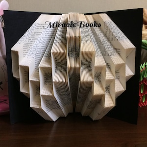May include: A folded book art sculpture featuring intricate page pleats creating a geometric pattern. The pages are cream-coloured with black printed text. The text Miracle Books is visible in black script. The sculpture is mounted on a black backing.