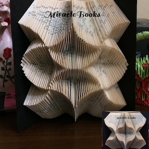 May include: A folded book art sculpture featuring intricate page folds that create a geometric star-like pattern. The pages are cream-coloured with visible black text. The text "Miracle Books" is printed in black across the centre of the sculpture.