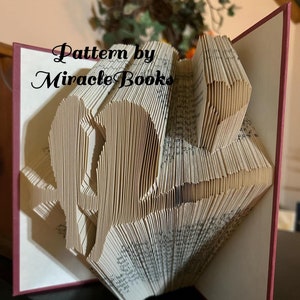 May include: A book art sculpture of a bird in flight, made by folding the pages of a book. The book is bound in a burgundy cover and the sculpture is titled "Pattern by Miracle Books".