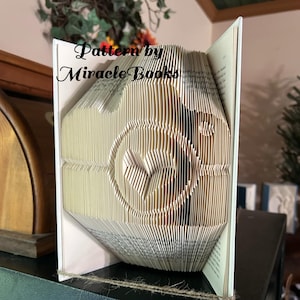 May include: A book folded into the shape of a camera. The book is standing on a dark green surface. The text "Pattern by Miracle Books" is visible on the front of the book.