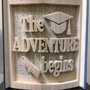 May include: A book art sculpture with the words "The Adventure Begins" in a 3D design. The sculpture also includes a graduation cap and a scroll.