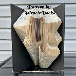 May include: A folded book art piece with the text "Pattern by Miracle Books". The book is folded into a geometric design, set within a black frame. The pages are beige, and the frame is black. A twine is wrapped around the base.