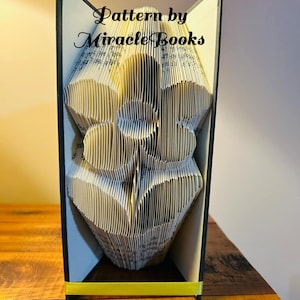 May include: A book art sculpture of a flower, folded from the pages of a book. The book is bound in black with a yellow ribbon. The sculpture is titled "Pattern by Miracle Books".
