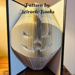 Peut inclure: Un livre plié en une sculpture 3D d'un visage. Le livre est ouvert et les pages sont pliées pour créer la forme d'un visage. Le livre est posé sur une table en bois. Le texte "Pattern by Miracle Books" est visible en haut de l'image.