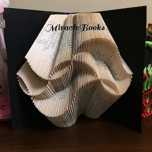 Puede incluir: Una escultura de libro plegado con páginas intrincadamente dobladas para formar un patrón geométrico, sobre un fondo negro. El texto Miracle Books está impreso en negro en la parte superior central de las páginas de papel color crema.