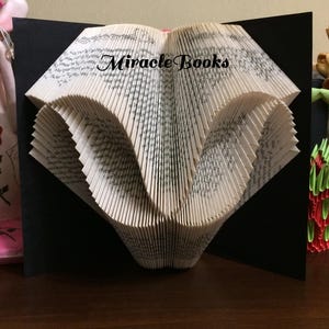 Könnte beinhalten: Eine Buchfaltkunst-Skulptur mit aufwendig gefalteten Seiten, die eine geometrische Form bilden, vor einem schwarzen Hintergrund. Der Text Miracle Books ist in Schwarz über die Mitte der weißen Papierseiten gedruckt, die sichtbaren schwarzen Text zeigen.