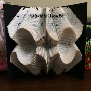 May include: A folded book sculpture featuring pages intricately creased to form a symmetrical geometric pattern. The centre of the sculpture displays the text "Miracle Books" in a black serif font, set against a solid black background panel.