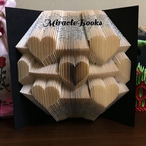 Puede incluir: Una escultura de libro plegado que presenta múltiples formas de corazón creadas a partir de las páginas de un libro. La escultura está montada sobre un fondo negro y muestra el texto Miracle Books en letras negras en la parte superior central.