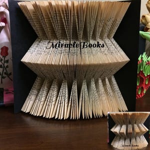 May include: A folded book art sculpture featuring pages arranged in a symmetrical hourglass pattern against a black backing. The text Miracle Books is printed in black script across the centre of the cream-coloured paper pages, which are folded to create a textured, pleated appearance.