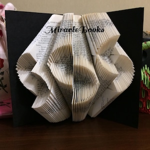 May include: A folded book art sculpture featuring pages intricately creased to form a geometric shape, set against a black background. The text "Miracle Books" is printed in black across the top centre of the white paper pages with visible text.