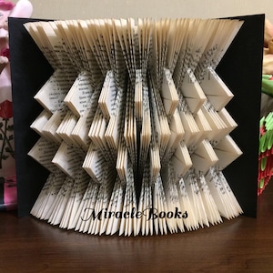 May include: A folded book art sculpture featuring pages intricately creased into a geometric, accordion-like pattern. The centre of the sculpture displays the text Miracle Books in a black script font against the white paper pages.