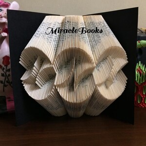 Puede incluir: Una escultura de libro plegado con páginas de color crema con pliegues simétricos e intrincados que crean un patrón geométrico. El texto "Miracle Books" está impreso en negro en la parte superior central de las páginas, sobre un fondo negro.