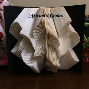 May include: A folded book art sculpture featuring cream-coloured pages arranged in a geometric pattern against a black background. The text Miracle Books is printed in black ink on the top centre of the folded pages, showcasing intricate paper folding.