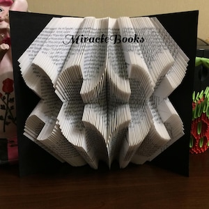 Peut inclure: Sculpture de livre plié avec le texte "Miracle Books" sur fond noir. Les pages sont pliées de manière complexe pour créer un design en trois dimensions. Le livre est posé sur une surface en bois.