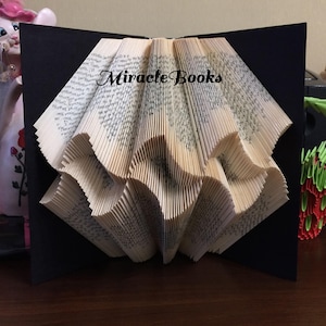May include: A unique book art creation featuring folded pages arranged in a fan shape. The book is displayed against a black backdrop, with the words "Miracle Books" printed on the pages. The folded pages create a textured, layered effect.