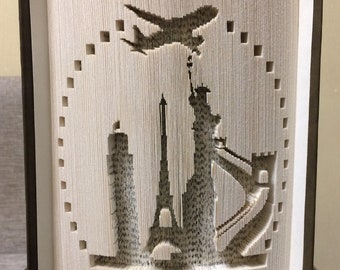May include: A book with pages folded into a 3D sculpture of the Statue of Liberty, the Eiffel Tower, and a skyscraper. An aeroplane flies above the city skyline. The book is titled "Miracle Books".