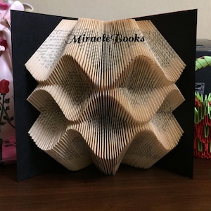 Op de afbeelding: Een handgemaakte papieren sculptuur gemaakt van gevouwen pagina's van een boek, in een geometrisch ontwerp. De sculptuur wordt gepresenteerd tegen een zwarte achtergrond, met de tekst "Miracle Books" zichtbaar.