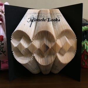 Op de afbeelding: Een gevouwen boekkunstsculptuur met ingewikkelde geometrische patronen gemaakt van individuele papieren pagina's. Het kunstwerk staat tegen een zwarte achtergrond en toont de tekst Miracle Books in zwarte letters in het midden bovenaan.