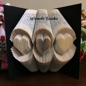 Op de afbeelding: Een gevouwen boekkunstsculptuur met drie hartvormen die zijn gecreëerd door de pagina's nauwkeurig te vouwen. De tekst "Miracle Books" is in het zwart gedrukt over het midden bovenaan de witte papieren pagina's tegen een zwarte achtergrond.