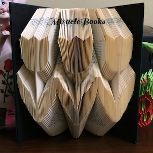 Op de afbeelding: Een gevouwen boeksculptuur met de tekst "Miracle Books" op de pagina's. De pagina's zijn gevouwen om een geometrisch ontwerp te creëren. Het boek staat tegen een donkere achtergrond.