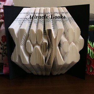 May include: A folded book art piece with the letters "XOX" cut into the pages. The book is open, revealing the folded pages in a fan-like shape. The text "Miracle Books" is printed on the top of the pages. The background is black.