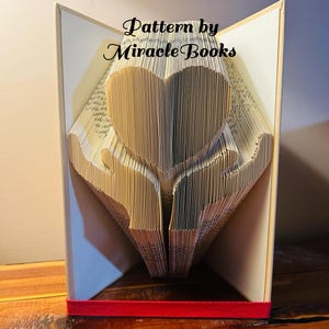 Könnte beinhalten: Ein Buch, das zu einem Herz gefaltet ist, mit Händen, die sich nach außen strecken. Das Buch ist geöffnet und der Rücken ist sichtbar. Das Buch liegt auf einer Holzoberfläche. Der Text "Pattern by Miracle Books" ist auf dem vorderen Umschlag sichtbar.